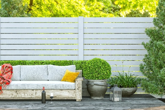 Gemütliche Terrasse mit Sofa, Pflanzen und einem weißen Sichtschutzzaun im Hintergrund.
